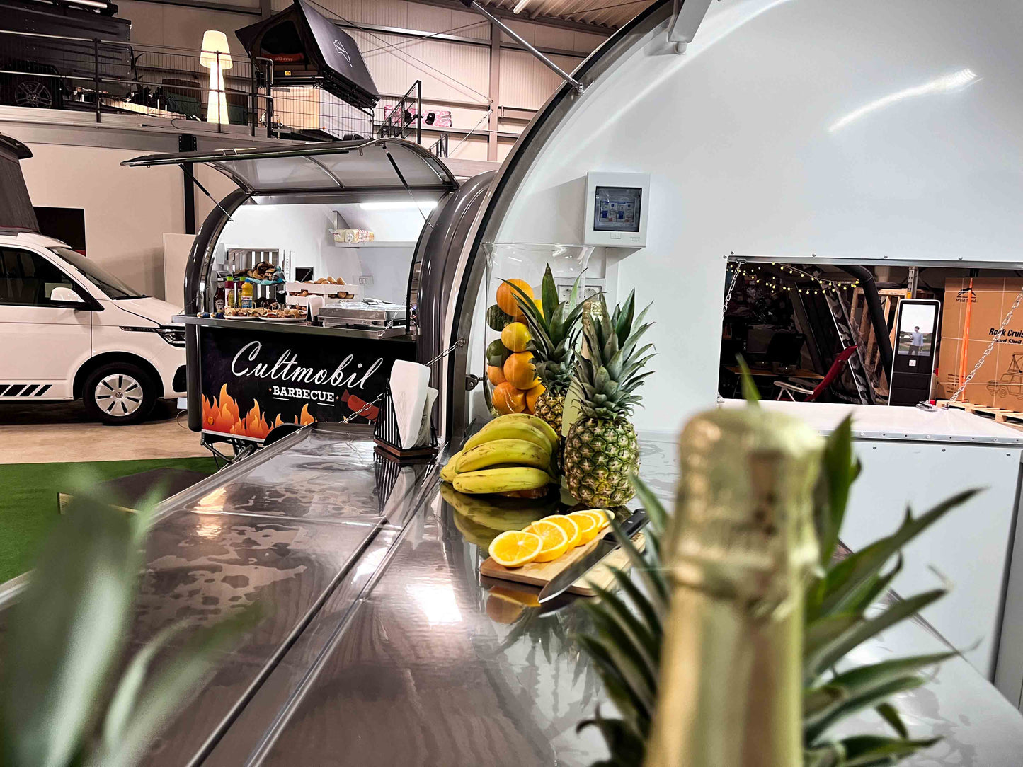 Innenansicht eines Foodtrailer mit Blick auf die Theke vorne. Die Theke ist dekoriert mit Obst. Im Hintergrund ist ein weiterer Verkaufsanhänger zu sehen. 