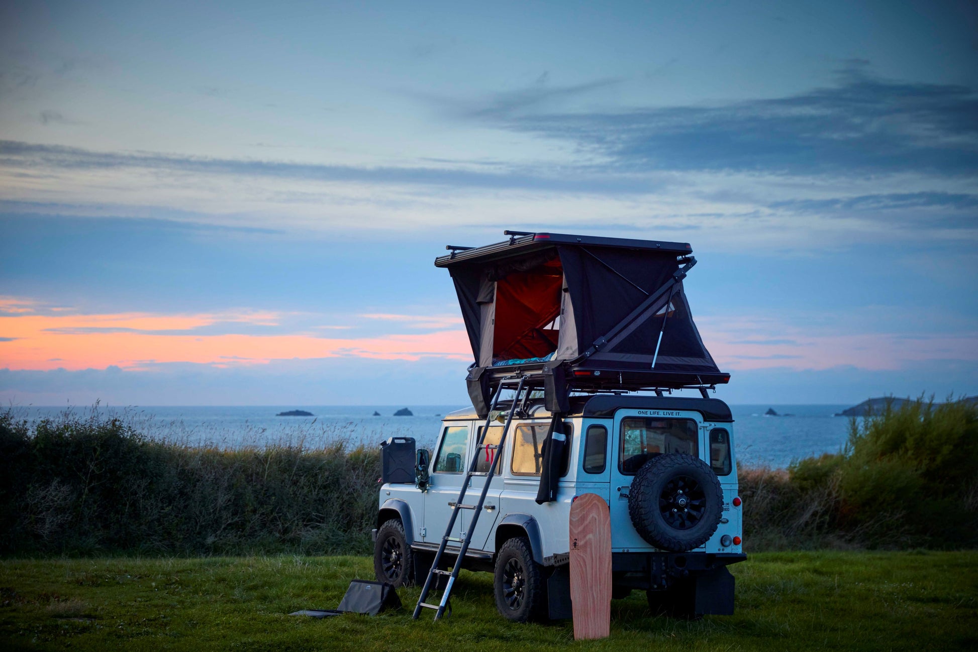 Dachzelt für Auto und Minicamper, 2 Personen, Hartschale mit Z-förmigem Bügel, auf einem Jeep montiert, stehend auf einem Rasenstreifen vor dem Strand.