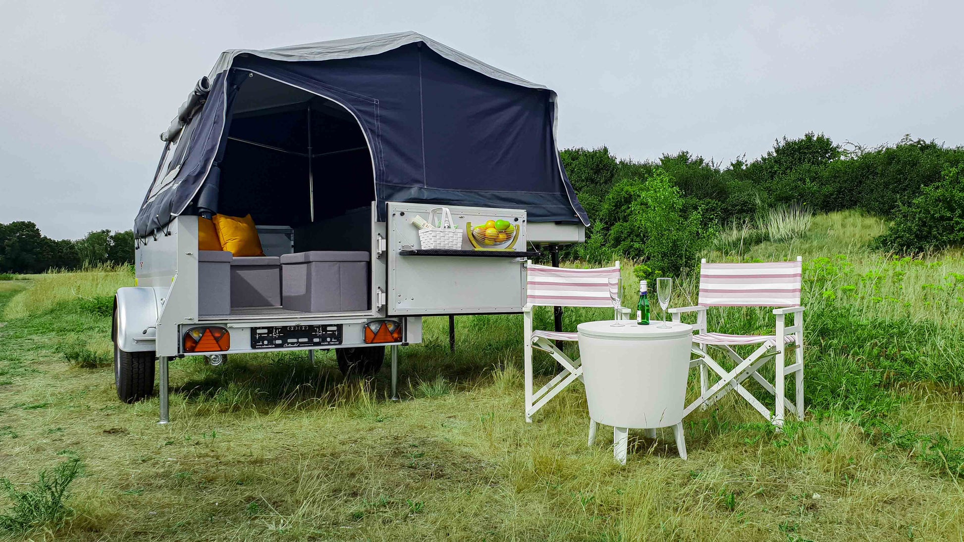 Blick auf einen vollständig aufgebauten kleinen Zeltanhänger mit Campingstühlen, Tisch und kleiner Küche
