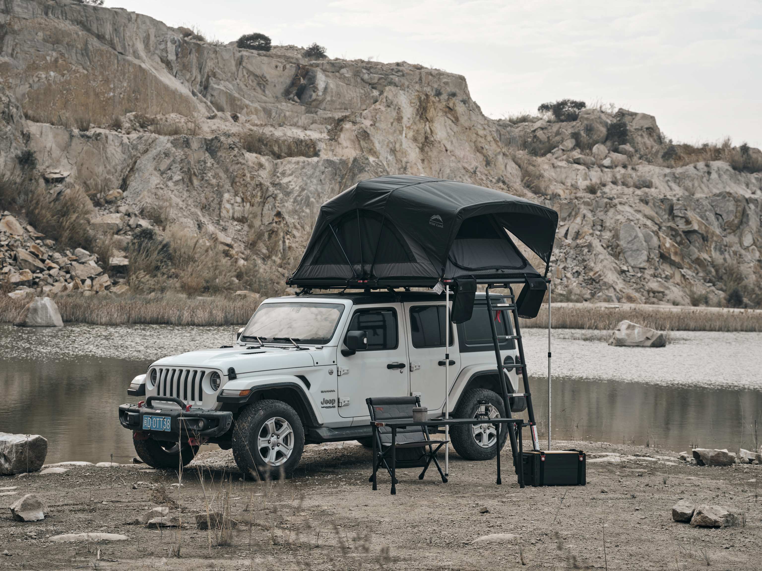 Dachzelt aufgebaut auf einem Jeep vor Bergen mit Campingstuhl und Tisch. 